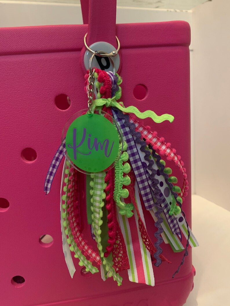 Pode incluir: Uma bolsa tote rosa com um chaveiro decorativo. O chaveiro apresenta uma etiqueta circular verde com o nome "Kim" em escrita cursiva e fitas coloridas em tons de rosa, roxo, verde e branco. A bolsa tem uma al&ccedil;a e um anel prateado.