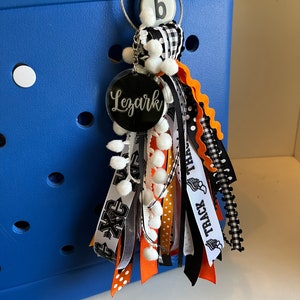 May include: A black and white tassel keychain with a round, black acrylic charm that says "Lezark" in white lettering. The tassel is made of various ribbons, including black and white checked ribbon, black and white polka dot ribbon, orange and white ribbon, and black ribbon with white text that says "Track Team". The keychain is attached to a blue tote bag.