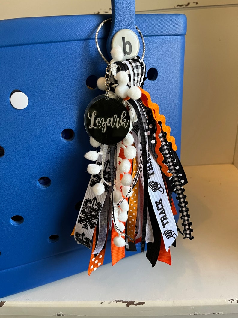 May include: A black and white tassel keychain with a round, black acrylic charm that says "Lezark" in white lettering. The tassel is made of various ribbons, including black and white checked ribbon, black and white polka dot ribbon, orange and white ribbon, and black ribbon with white text that says "Track Team". The keychain is attached to a blue tote bag.
