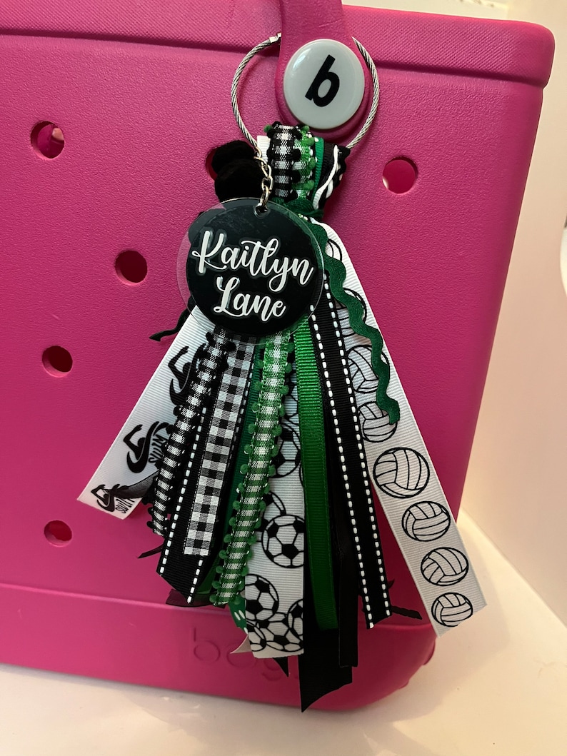 May include: A black and white checked ribbon with green and black ribbons, a white ribbon with black footballs, and a white ribbon with black volleyballs. The ribbons are attached to a round black tag with the name "Kaitlyn Lane" written in white. The tag is attached to a pink bag with a white button that says "b".