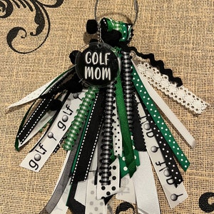 May include: A black and green golf-themed keyring with a round tag that says "GOLF MOM" in white lettering. The keyring has multiple ribbons in black, white, and green with golf ball and club designs.