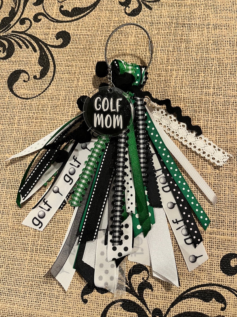 May include: A black and green golf-themed keyring with a round tag that says "GOLF MOM" in white lettering. The keyring has multiple ribbons in black, white, and green with golf ball and club designs.