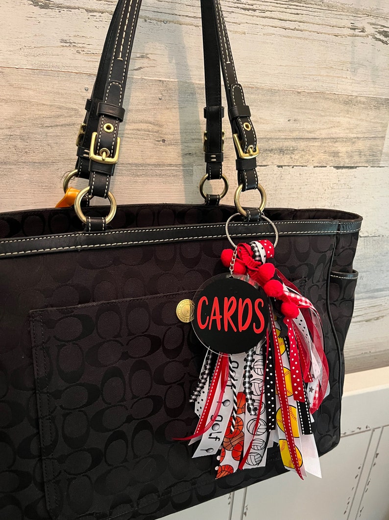 May include: Black tote bag with a black and white patterned design. The bag has a gold zip and a black leather strap. A black and red keychain with the word "CARDS" is attached to the bag.