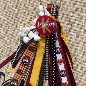 May include: A red and gold keychain with a round charm that says "Kristen" and a variety of ribbons in red, black, white, and gold. The ribbons have a checkered pattern, polka dots, and a logo with a mascot.