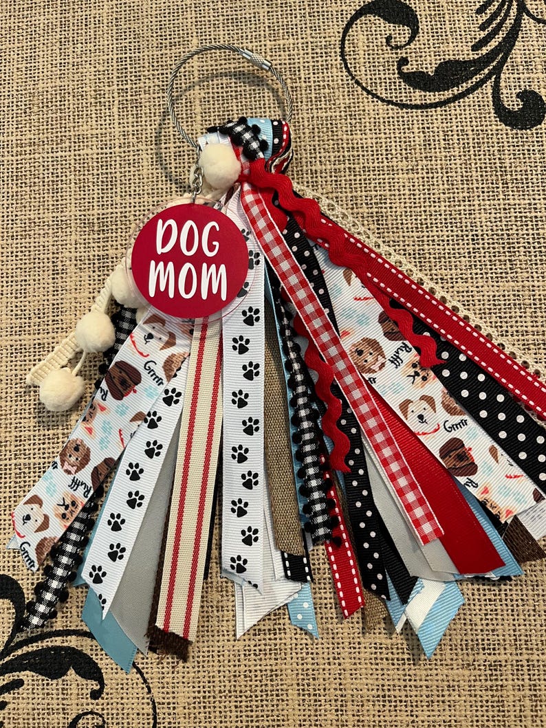 May include: A keychain with a red disc bearing the text "DOG MOM". It has a variety of ribbons with dog and paw print patterns in red, black, white, and blue. The keychain also includes a metal ring and small pom-poms for decoration.