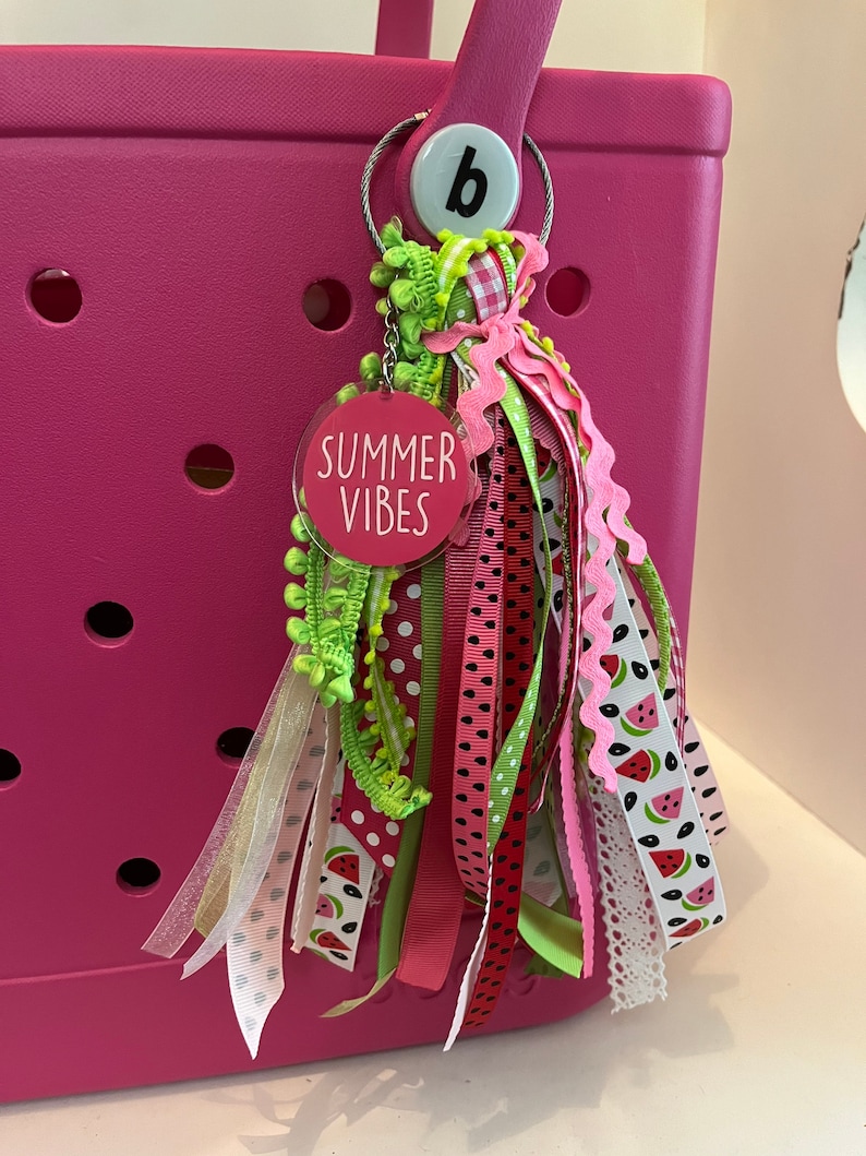 May include: A pink tote bag with a watermelon-themed keychain. The keychain has a round tag that says "Summer Vibes" and is attached to a ring with a letter "b" on it. The keychain has a variety of colorful ribbons and trims.