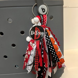 May include: A grey tote bag with a black and white, red and orange tassel keychain. The keychain has a clear circle charm with the text "De Long" and a red letter "D".
