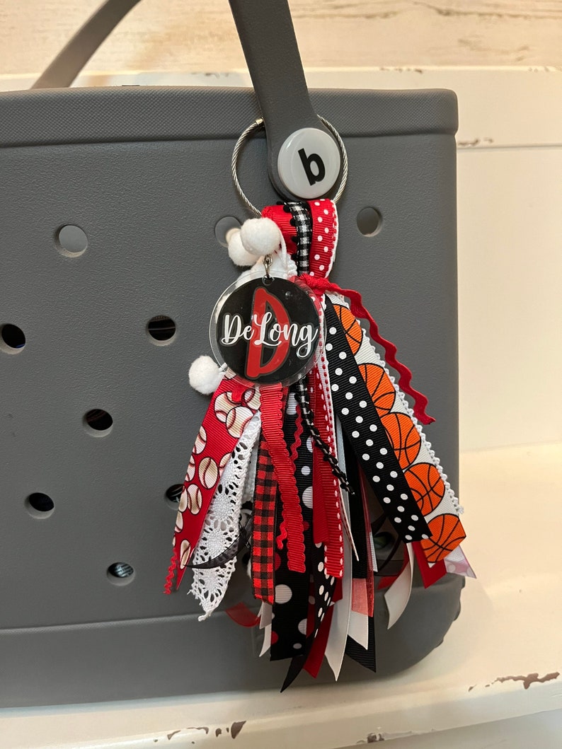 May include: A grey tote bag with a black and white, red and orange tassel keychain. The keychain has a clear circle charm with the text "De Long" and a red letter "D".