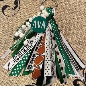 May include: Green, white and black cheerleading keychain with a round green acrylic charm that says "AVA" and a variety of ribbons with different patterns, including polka dots, stripes and checkered.