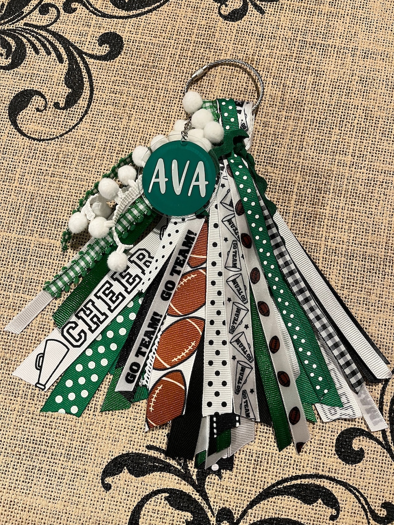 May include: Green, white and black cheerleading keychain with a round green acrylic charm that says "AVA" and a variety of ribbons with different patterns, including polka dots, stripes and checkered.
