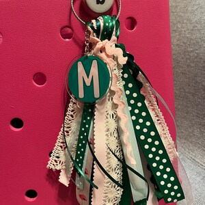 May include: A green and white polka dot ribbon charm with a green circle charm with the letter "M" in pink. The charm is attached to a pink bag with a silver keychain.