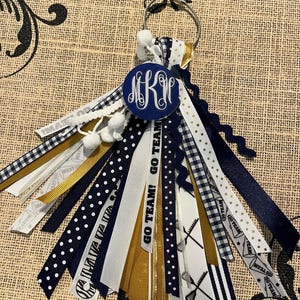 May include: A navy blue and gold lacrosse keychain with a white pom-pom and a monogrammed circle charm. The keychain has multiple ribbons with different patterns, including polka dots, stripes and plaid. The text "GO TEAM! GO TEAM!" is printed on one of the ribbons.