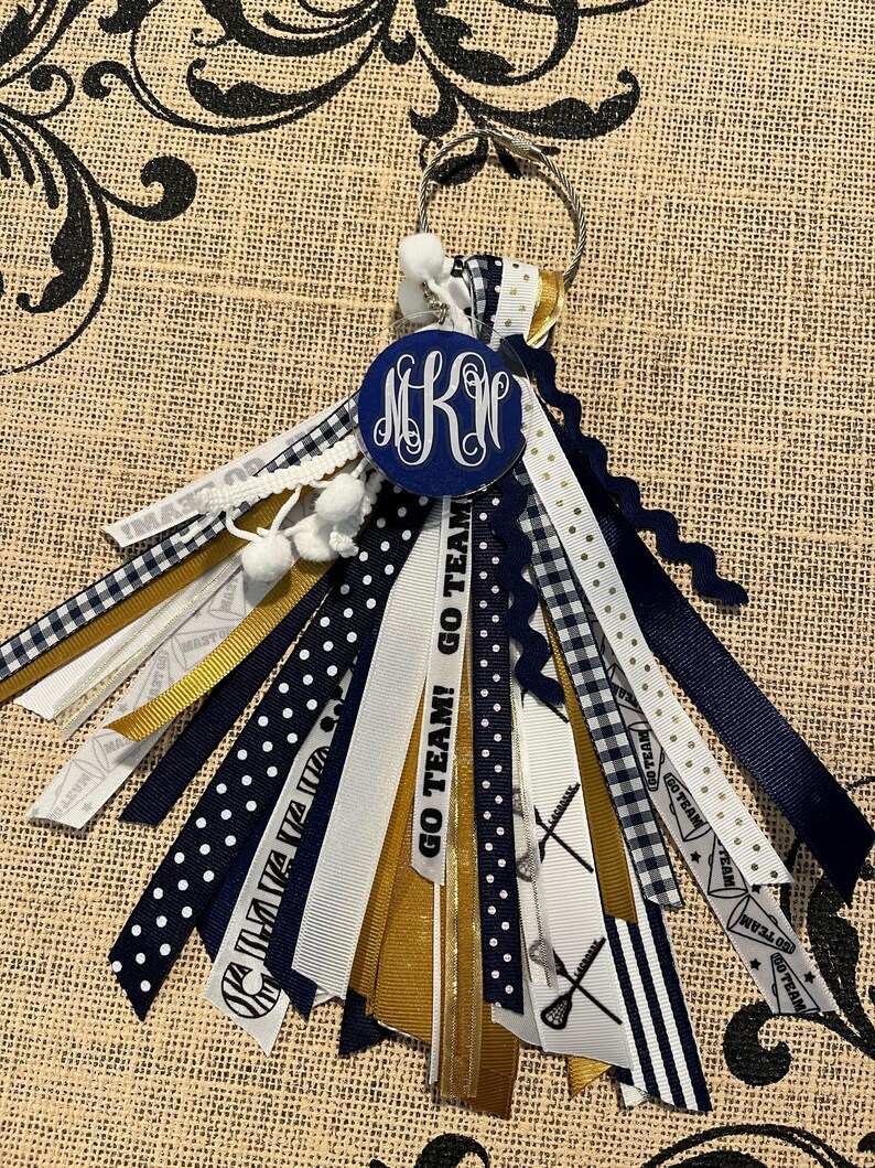 May include: A navy blue and gold lacrosse keychain with a white pom-pom and a monogrammed circle charm. The keychain has multiple ribbons with different patterns, including polka dots, stripes and plaid. The text "GO TEAM! GO TEAM!" is printed on one of the ribbons.