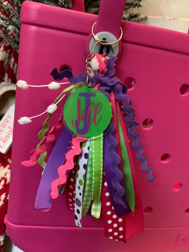 Pode incluir: Um porta-chaves personalizado com uma etiqueta circular verde que diz "Jill" em escrita rosa. O porta-chaves apresenta uma variedade de fitas coloridas em tons de rosa, roxo e verde, presas a um porta-chaves dourado. O porta-chaves est&aacute; preso a uma sacola rosa brilhante.