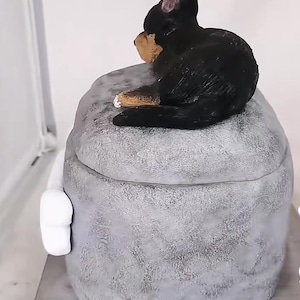 May include: A decorative, grey, textured box with a lid, topped with a black and tan dog figurine. The box has a white handle on the side and sits on a wooden base. The dog is in a seated position.