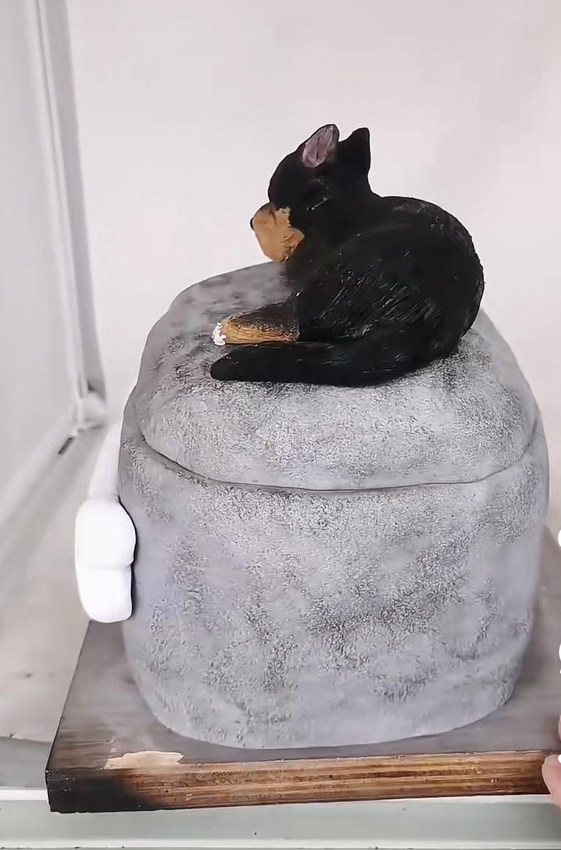 May include: A decorative, grey, textured box with a lid, topped with a black and tan dog figurine. The box has a white handle on the side and sits on a wooden base. The dog is in a seated position.