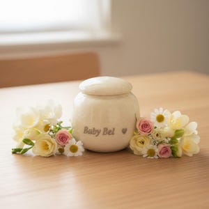 May include: A cream ceramic urn with a lid, inscribed with "Baby Bel" and a heart symbol. The urn is surrounded by arrangements of white, yellow, and pink flowers, set on a light wooden surface.