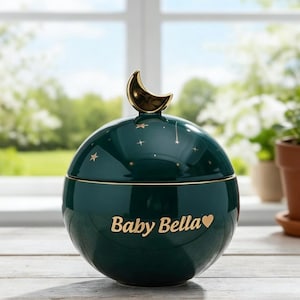 May include: A dark teal ceramic jar with a gold crescent moon handle. The jar is round with gold stars and the words "Baby Bella" in gold script. A heart symbol follows the name.