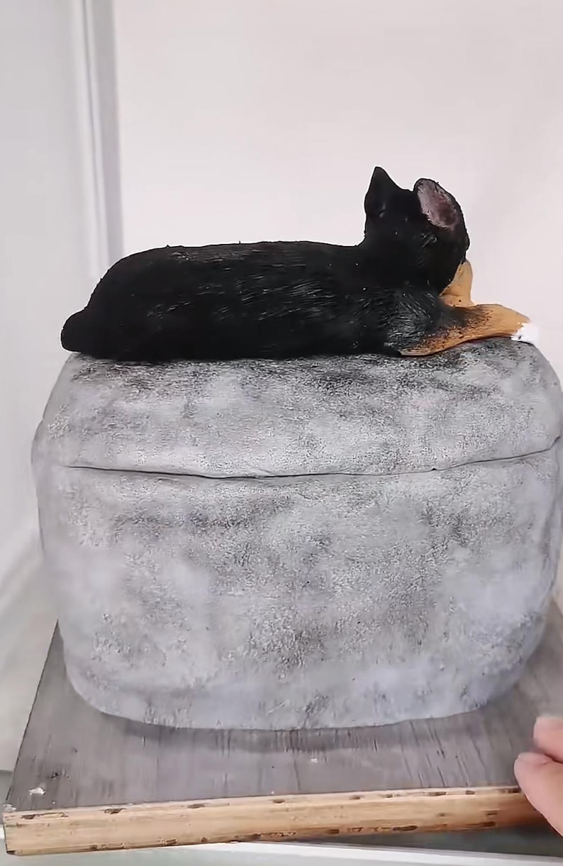 May include: A decorative, grey, stone-like box with a black and tan dog figurine resting on top. The dog is lying down, with its head and ears visible. The box has a rounded shape and a textured surface.