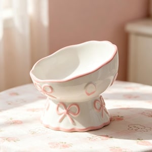 May include: A white ceramic pet bowl with a scalloped rim and pink trim. The bowl features pink bow accents around the base and rim. The bowl is elevated on a pedestal base, creating a stylish and functional design.