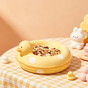 May include: A yellow ceramic duck-shaped pet bowl with white polka dots, filled with birdseed. The bowl has a duck head on one side. A small yellow duck figurine and other decorative figurines are nearby. The bowl sits on a yellow and white checkered tablecloth.