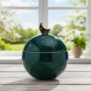 Può includere: Un barattolo in ceramica verde scuro con una maniglia a forma di mezzaluna dorata. Il barattolo ha una forma sferica con dettagli di stelle dorate. Il barattolo è appoggiato su una superficie di legno davanti a una finestra.