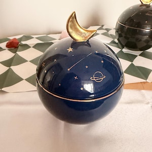 Peut inclure: Boîte à bijoux sphérique en céramique bleu marine avec une poignée en forme de croissant de lune dorée. Le coffret présente des étoiles dorées et un motif de planète. Une deuxième boîte noire avec une lune dorée est visible.