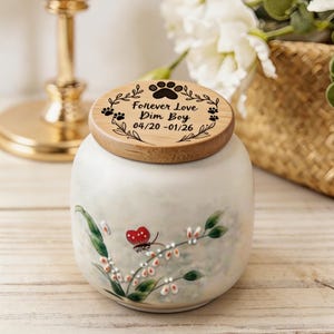Puede incluir: Urna para mascotas de cerámica blanca con tapa de madera. La tapa está grabada con huellas de patas, las palabras "Forever Love Dim Boy 04/20 - 01/26" y hojas decorativas. La urna está decorada con una mariposa y motivos florales.