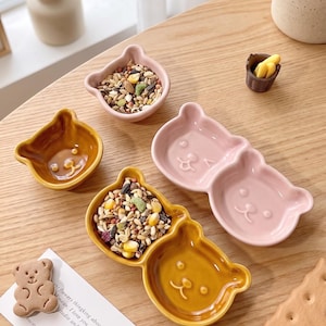 以下が含まれることがあります： ピンクと黄色のクマの形をした陶器の食器のコレクション。一部は種で満たされています。小さなクマの形をしたクッキーとテキストが書かれたカードも表示されています。食器は木製の表面に置かれています。