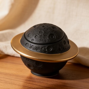 Puede incluir: Una caja de baratijas de cerámica negra con forma de planeta con un anillo dorado. La superficie del planeta está texturizada con cráteres. La caja está sobre una superficie de madera.