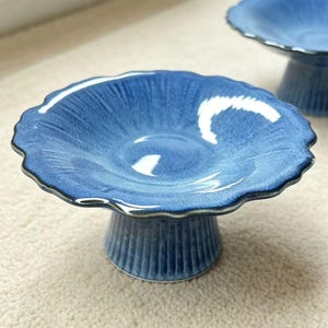 May include: Two blue ceramic pedestal bowls with scalloped edges. The bowls have a textured surface and a dark blue rim. The pedestal base is a darker blue with vertical grooves. The bowls are on a light-colored surface.