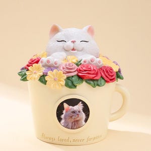 May include: A cream-colored cat figurine in a teacup-shaped container, surrounded by colorful flowers. The cat is white with closed eyes and a smiling expression. The teacup has a picture of a cat and the text "Always loved, never forgotten."