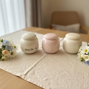 Könnte beinhalten: Drei Keramikurnen mit Deckeln, in Weiß und Rosa, stehen auf einer beigen Tischdecke. Die Urnen sind von Sträußen aus weißen, gelben und blauen Blumen umgeben. Eine Urne trägt den Text "Baby Bel".