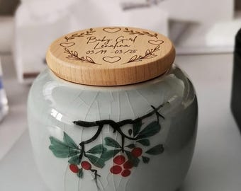 May include: A ceramic urn with a light blue glaze and a wooden lid. The lid is engraved with the words "Baby Girl Lenafina" and dates. The urn is decorated with a painted floral design featuring red berries and green leaves.