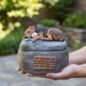 May include: A memorial pet urn with a sleeping dog figurine on top. The dog is brown and black, resting on a gray stone-like urn. A small ball and a blue toy are next to the dog. The urn has a plaque with text.
