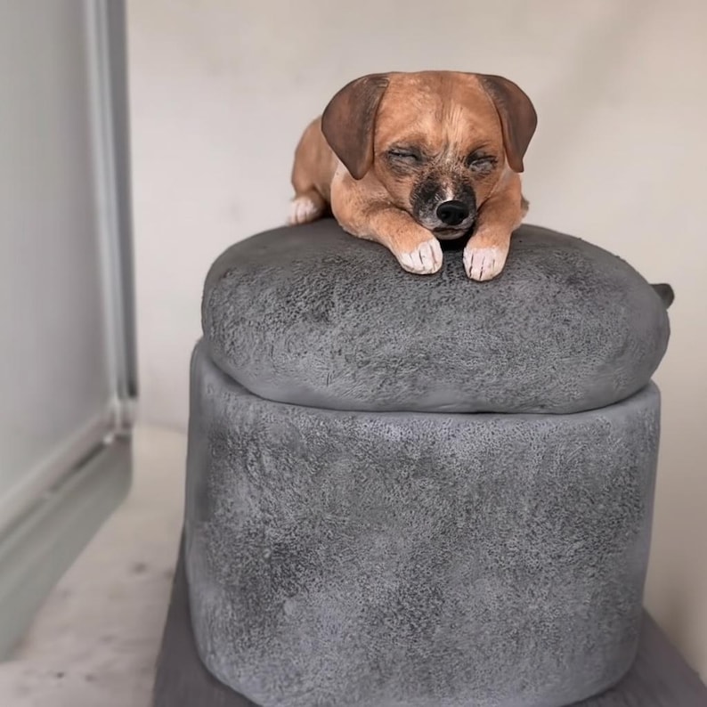 May include: A brown and white dog figurine with closed eyes rests on a grey, stone-like container. The dog has brown ears and a black nose. The container has a rounded top and a cylindrical base, suggesting a decorative storage item.