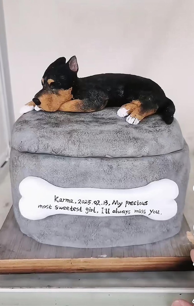 May include: A grey pet urn with a sculpted dog figurine on top. The urn has a bone-shaped plaque with the inscription "Karma, 2025.02.13, My precious most sweetest girl, I'll always miss You."