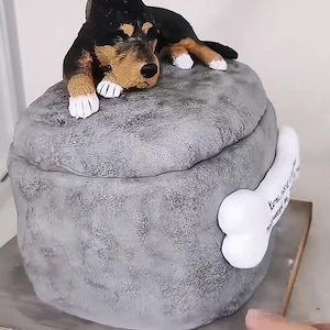 May include: A grey, stone-like sculpture of a dog lying on a rounded base. The dog is black and tan, with white paws. A white bone with text is attached to the side of the base. The sculpture is on a wooden base.