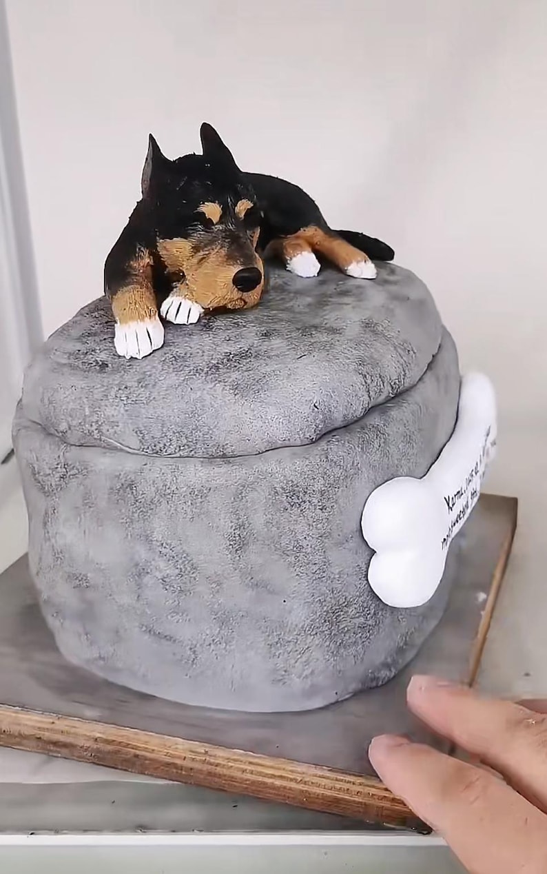 May include: A grey, stone-like sculpture of a dog lying on a rounded base. The dog is black and tan, with white paws. A white bone with text is attached to the side of the base. The sculpture is on a wooden base.