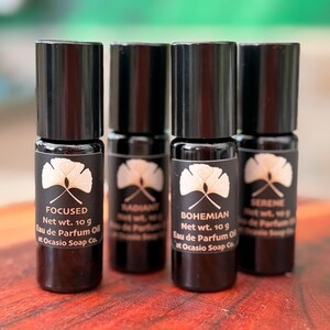 May include: Four small, dark-colored perfume bottles with black caps. Each bottle has a label with a white floral design and the words "FOCUSED", "RADIANT", "BOHEMIAN", and "SERENE". The labels also include "Net wt. 10 g Eau de Parfum Oil".