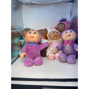 May include: A collection of six dolls with round faces and soft bodies, each wearing a different hooded onesie. The dolls feature outfits in shades of purple, white, pink, and orange, with some hoods designed as animal or mythical creature heads.