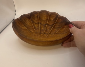 May include: A wooden, shell-shaped bowl with a natural wood grain finish. The bowl has a scalloped edge and a carved, fan-like pattern on the interior. The bowl is a warm brown color and appears to be made of a solid piece of wood.
