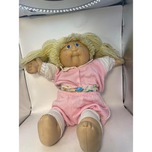 May include: A Cabbage Patch Kids doll with blonde yarn hair styled in pigtails, blue eyes, and a round face. The doll wears a pink short-sleeved top with a white collar and matching pink shorts over white leggings with thin grey stripes.