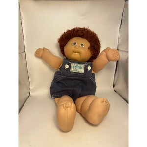 May include: A Cabbage Patch Kids doll with a round face, light skin, and short, curly auburn yarn hair. The doll wears dark blue denim overalls with white buttons and a rectangular patch on the chest featuring the Cabbage Patch Kids logo.