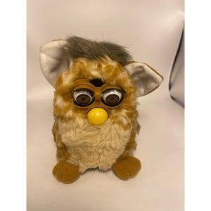 May include: A plush toy featuring a furry body with a mix of tan and white fur, a tuft of gray hair on top, and large plastic eyes with brown irises. The toy has prominent white-lined ears, a yellow beak, and two brown feet at the base.