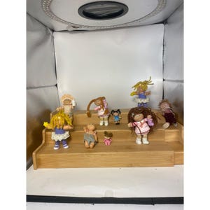 May include: A collection of nine small plastic dolls with yarn hair, featuring various outfits including dresses, skirts, and overalls. The dolls are arranged on a three-tiered wooden display stand, showcasing diverse hair colors, styles, and facial expressions.