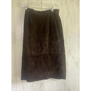 May include: A dark brown suede skirt hangs from a hanger. The skirt is knee-length and has a simple, classic design. The suede has a soft, textured appearance, and the color is a rich, earthy brown.
