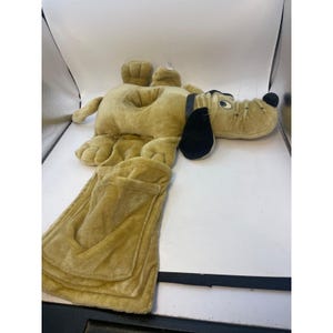 May include: A plush dog-shaped pillow in a light brown color. The dog has black ears, a black nose, and a long, rectangular pocket attached to its body. The pillow is designed to be used as a travel pillow or a decorative item.