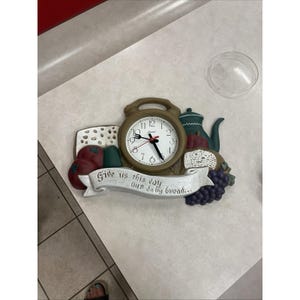 May include: A wall clock shaped like a kitchen scene with a central round clock face, a green teapot, red tomatoes, bread, and purple grapes. A white banner across the bottom features the text "Give us this day our daily bread..." in black.