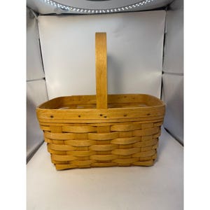 May include: A rectangular, light brown woven basket with a tall, vertical handle. The basket is made of woven wood strips, with a sturdy construction. The handle is also made of wood and is attached to the basket with small metal rivets. The basket is empty.